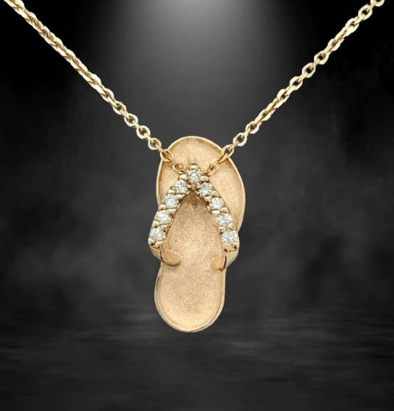 May include: Gold-toned pendant necklace featuring a flip-flop design. The flip-flop has a textured surface and is adorned with small, sparkling gemstones along the straps. The chain is delicate and complements the pendant's design.