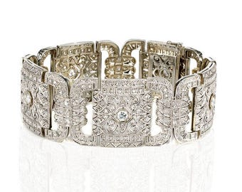 May include: An ornate silver bracelet with a rectangular design. The bracelet is encrusted with numerous small, sparkling stones, and features intricate filigree patterns. The clasp is visible, and the bracelet is displayed on a white surface.