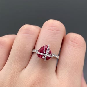May include: A heart-shaped ruby ring with a bow design. The ring features a large, faceted red gemstone at the center, accented by a delicate bow and a band encrusted with small, sparkling stones. The ring is set in a silver or white gold setting.
