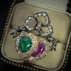 May include: An antique ring featuring a bow and two heart-shaped gemstones. One heart holds a green emerald, the other a pink ruby, both surrounded by diamonds. The ring is set in gold and silver, displayed in a vintage jewelry box.