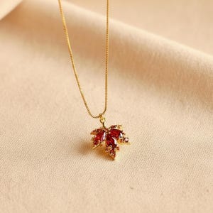 Op de afbeelding: Een goudkleurige ketting met een hanger in de vorm van een esdoornblad. Het blad is versierd met rode edelstenen. De ketting is fijn en hangt aan de hanger. De achtergrond is zacht en neutraal van kleur.