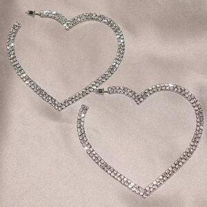 May include: A pair of heart-shaped hoop earrings. The earrings are encrusted with small, sparkling rhinestones. The earrings are silver-toned and have a post closure. The earrings are displayed on a light pink, satin-like surface.