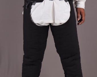 May include: Black quilted chaps featuring a wide waistband with two front buckle closures. The leg sections are constructed with vertical stitching and include side lacing details that run down the length of each leg, secured with black cord ties.