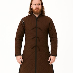 A man with a red beard wearing a long brown quilted gambeson coat with black trim and tie closures, standing against a white backdrop.