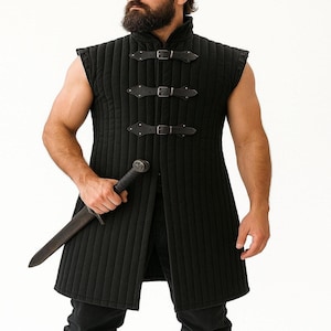 A bearded man wearing a black sleeveless quilted gambeson holds a dagger against a white background.