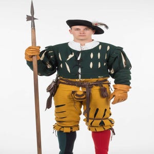 Könnte beinhalten: Historisches Kostüm mit dunkelgrünem Wams mit weißen Akzenten, gelber Hose und schwarzem Hut mit Feder. Das Outfit umfasst braune Lederhandschuhe, einen Gürtel mit Taschen und einen Speer. Die Hose hat rote und grüne Akzente.