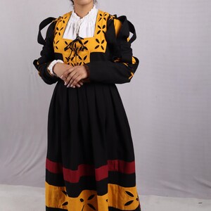 Pode incluir: Um vestido preto e amarelo com mangas compridas e saia rodada. O corpete apresenta um painel amarelo com desenhos recortados e uma gola branca. A saia tem listras horizontais amarelas e vermelhas. O vestido parece um traje hist&oacute;rico.