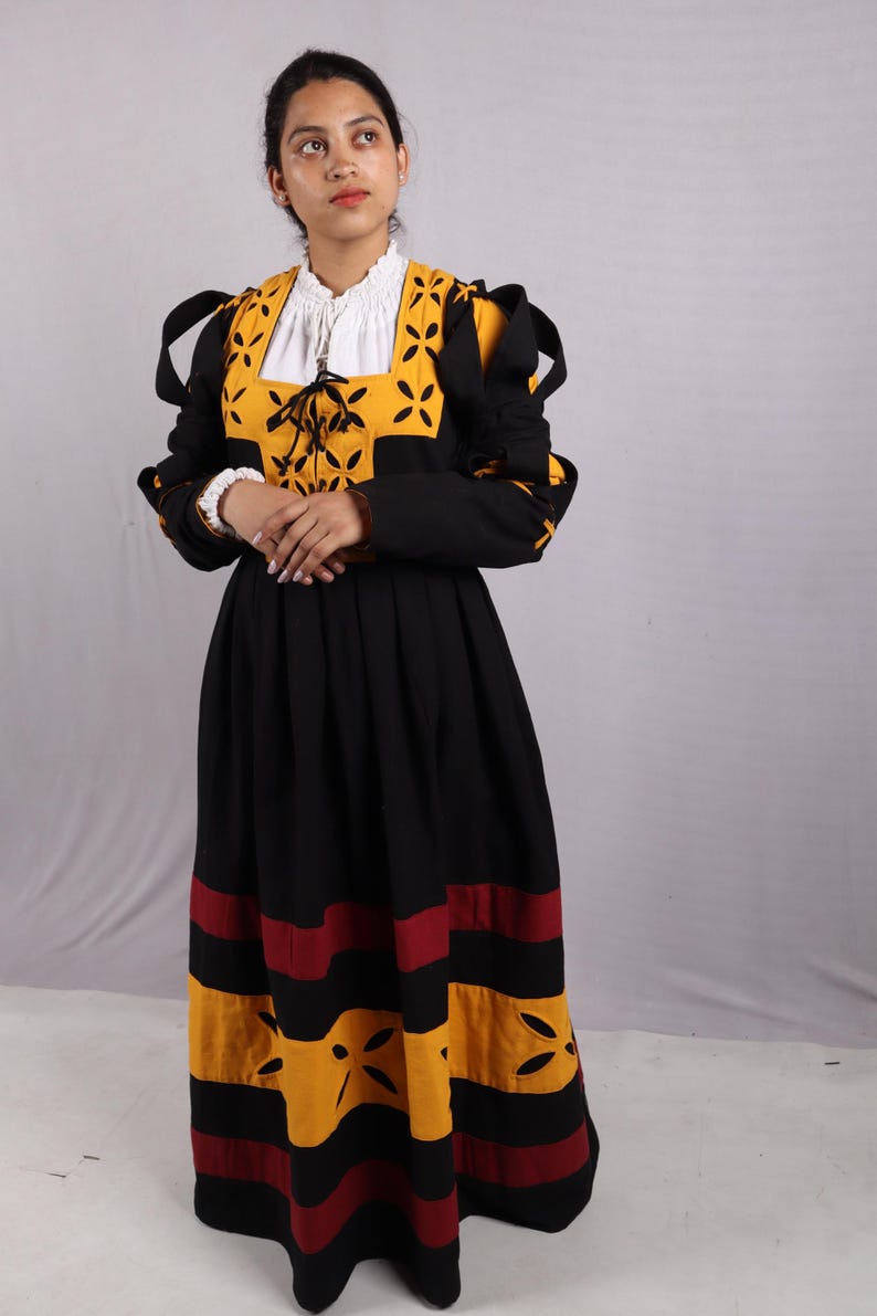 Pode incluir: Um vestido preto e amarelo com mangas compridas e saia rodada. O corpete apresenta um painel amarelo com desenhos recortados e uma gola branca. A saia tem listras horizontais amarelas e vermelhas. O vestido parece um traje hist&oacute;rico.