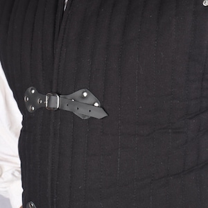 A close-up of the front of a black quilted gambeson featuring black leather straps with silver buckles fastened across the chest.