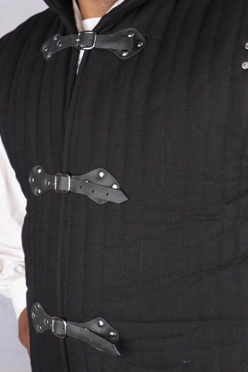 A close-up of the front of a black quilted gambeson featuring black leather straps with silver buckles fastened across the chest.