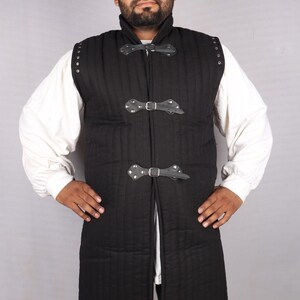 A bearded man wearing a black sleeveless quilted gambeson over a white shirt with his hands on his hips.