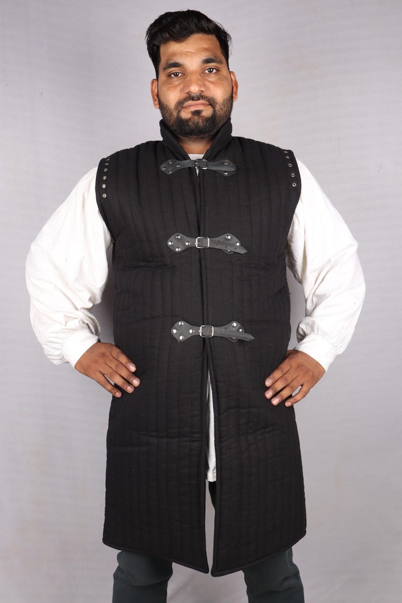 A bearded man wearing a black sleeveless quilted gambeson over a white shirt with his hands on his hips.