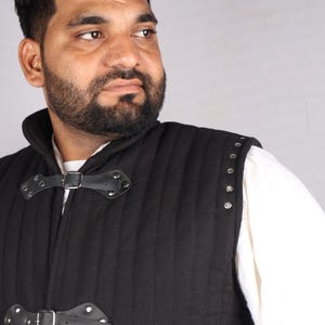 A close-up of a bearded man wearing a black quilted vest with leather straps and silver metal grommets along the shoulder.