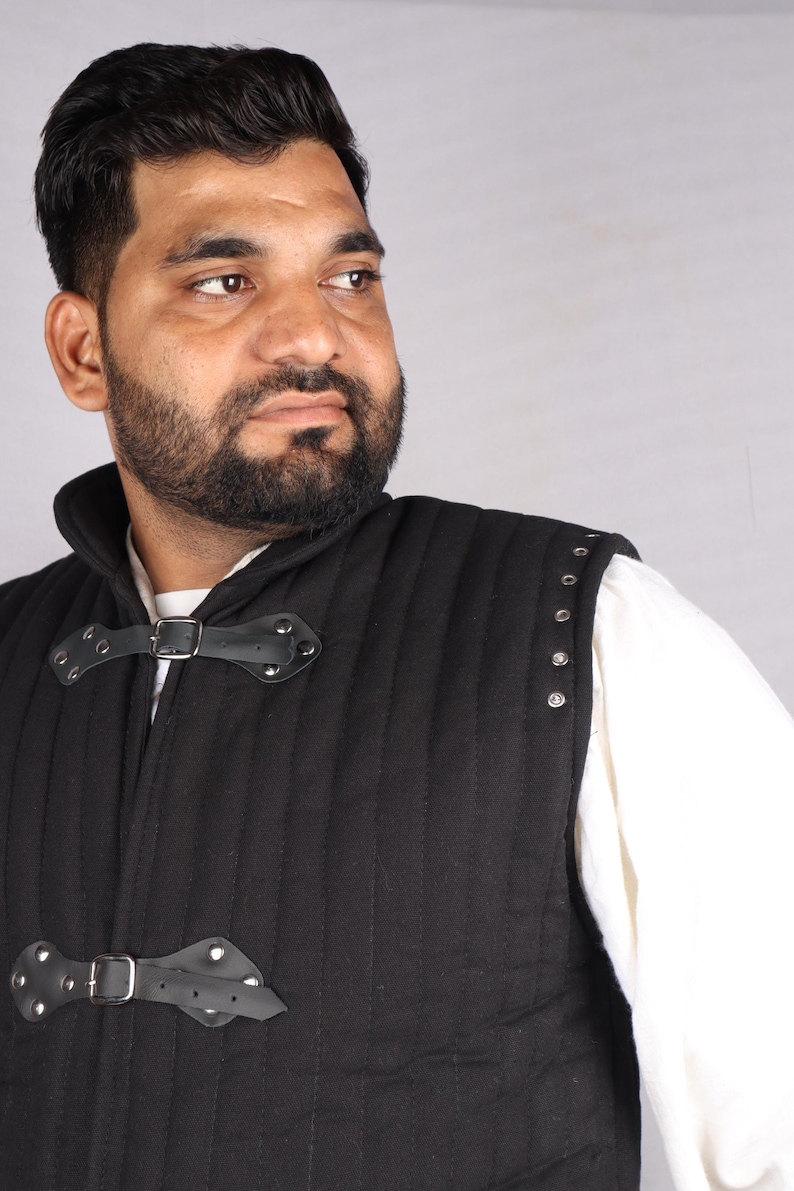 A close-up of a bearded man wearing a black quilted vest with leather straps and silver metal grommets along the shoulder.
