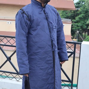 Full-body front view of a man wearing a long blue quilted gambeson coat with elaborate ties at the collar.