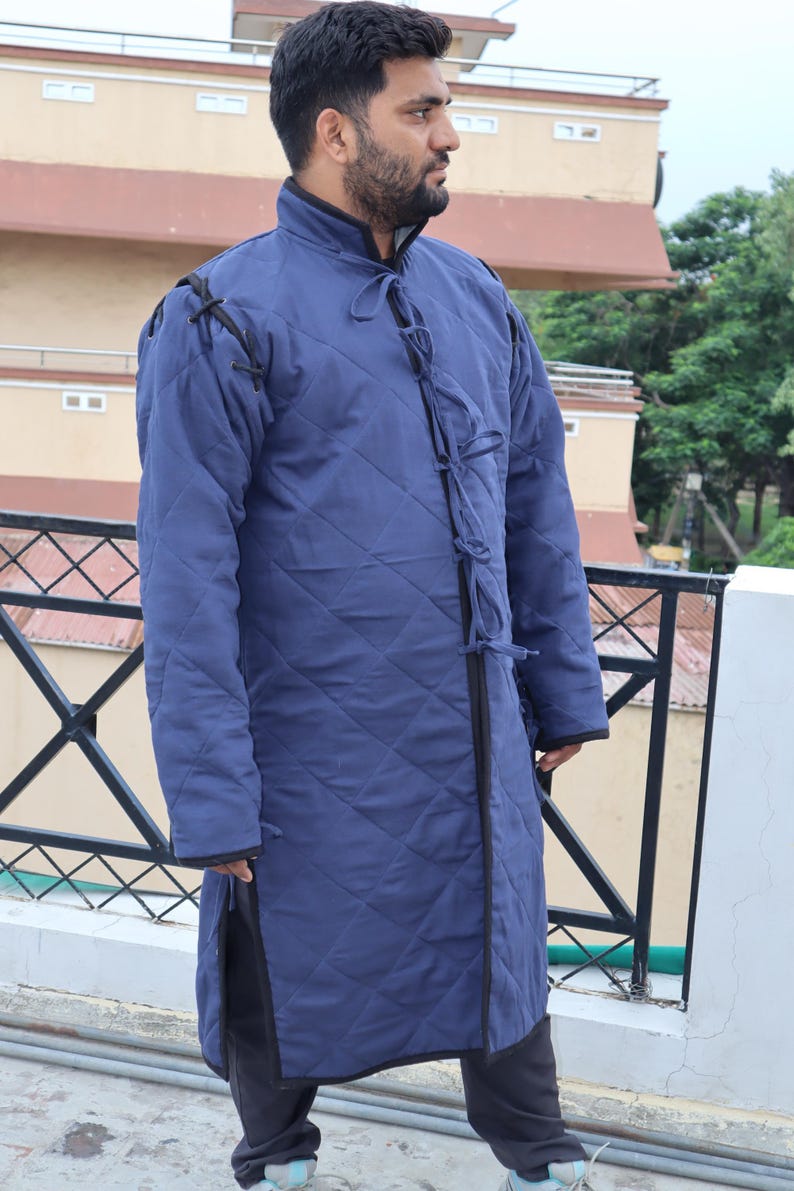 Full-body front view of a man wearing a long blue quilted gambeson coat with elaborate ties at the collar.