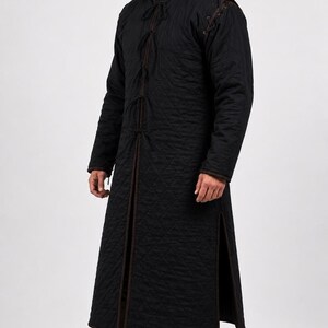 A man wearing a black quilted long gambeson coat shown in profile against a white backdrop.