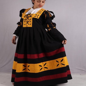 Pode incluir: Um vestido longo preto com detalhes amarelos e vermelhos. O corpete apresenta um painel amarelo com um design floral e uma gola branca. As mangas t&ecirc;m detalhes amarelos, e a saia tem listras horizontais amarelas e vermelhas com formas florais recortadas.