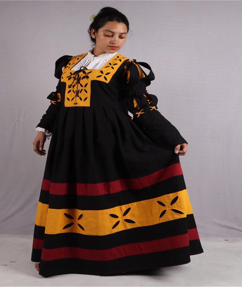 Pode incluir: Um vestido longo preto com detalhes amarelos e vermelhos. O corpete apresenta um painel amarelo com um design floral e uma gola branca. As mangas t&ecirc;m detalhes amarelos, e a saia tem listras horizontais amarelas e vermelhas com formas florais recortadas.