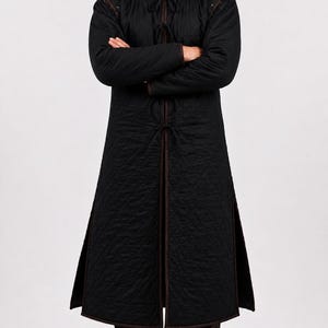 A man wearing a black quilted long gambeson coat standing with arms crossed against a white backdrop.