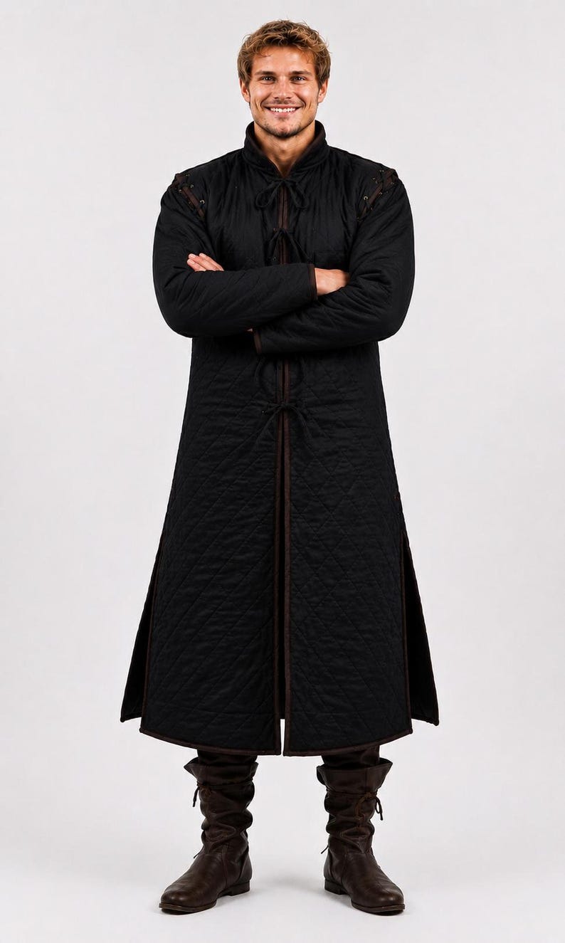 A man wearing a black quilted long gambeson coat standing with arms crossed against a white backdrop.