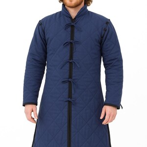A man with a beard wearing a long blue quilted gambeson coat with black trim and tie closures, standing against a white background.