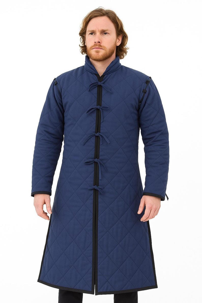 A man with a beard wearing a long blue quilted gambeson coat with black trim and tie closures, standing against a white background.