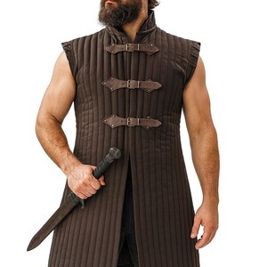 A bearded man wearing a brown sleeveless quilted gambeson and holding a dagger against a white background.