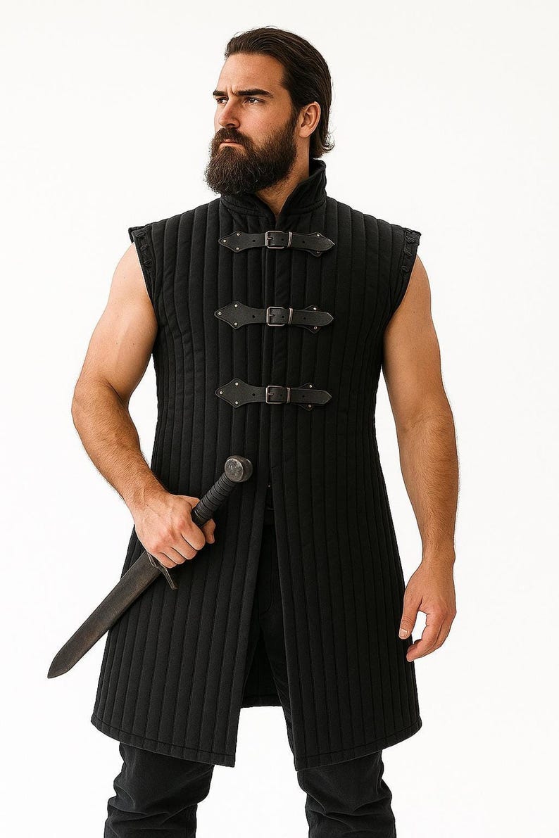 A bearded man wearing a black sleeveless quilted gambeson holding a dagger against a plain white background.
