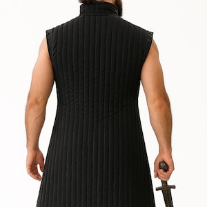 The back view of a bearded man with hair in a bun wearing a black quilted sleeveless gambeson holding a dagger.