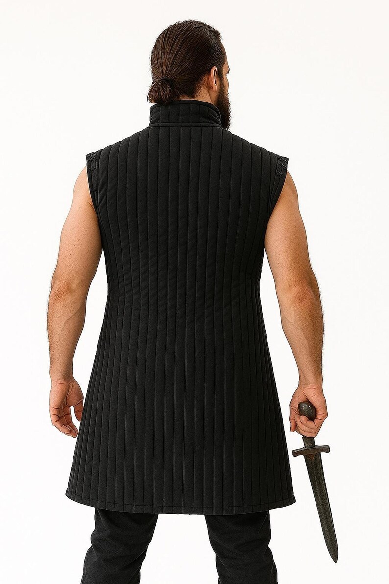 The back view of a bearded man with hair in a bun wearing a black quilted sleeveless gambeson holding a dagger.