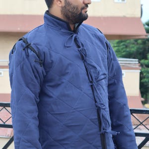 Close-up front view of a man wearing a blue quilted medieval coat with black shoulder lacing and multiple fabric ties down the chest.