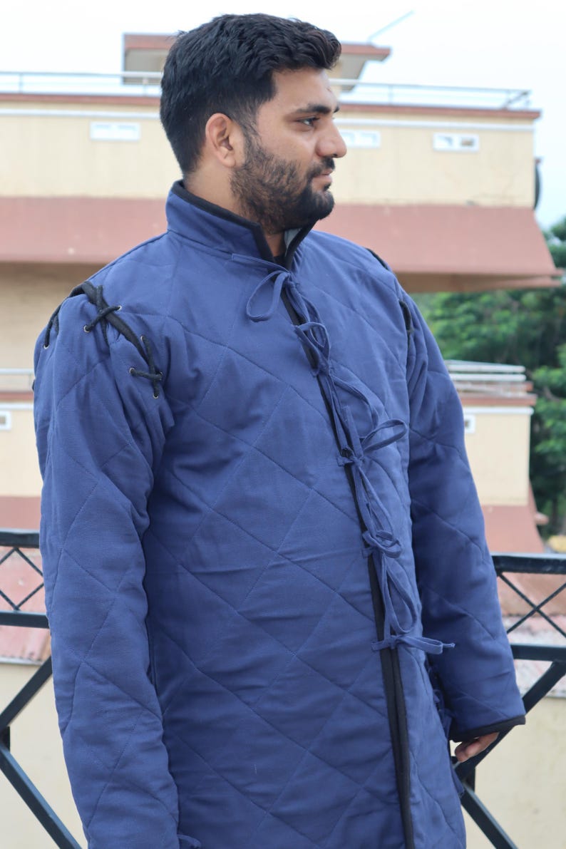 Close-up front view of a man wearing a blue quilted medieval coat with black shoulder lacing and multiple fabric ties down the chest.
