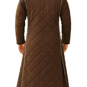 The back view of a person wearing a long brown quilted gambeson coat with yellow piping along the seams.