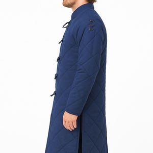 A male model wearing a long, blue quilted gambeson coat with a standing collar and front toggle closures, shown in profile against a plain white background.