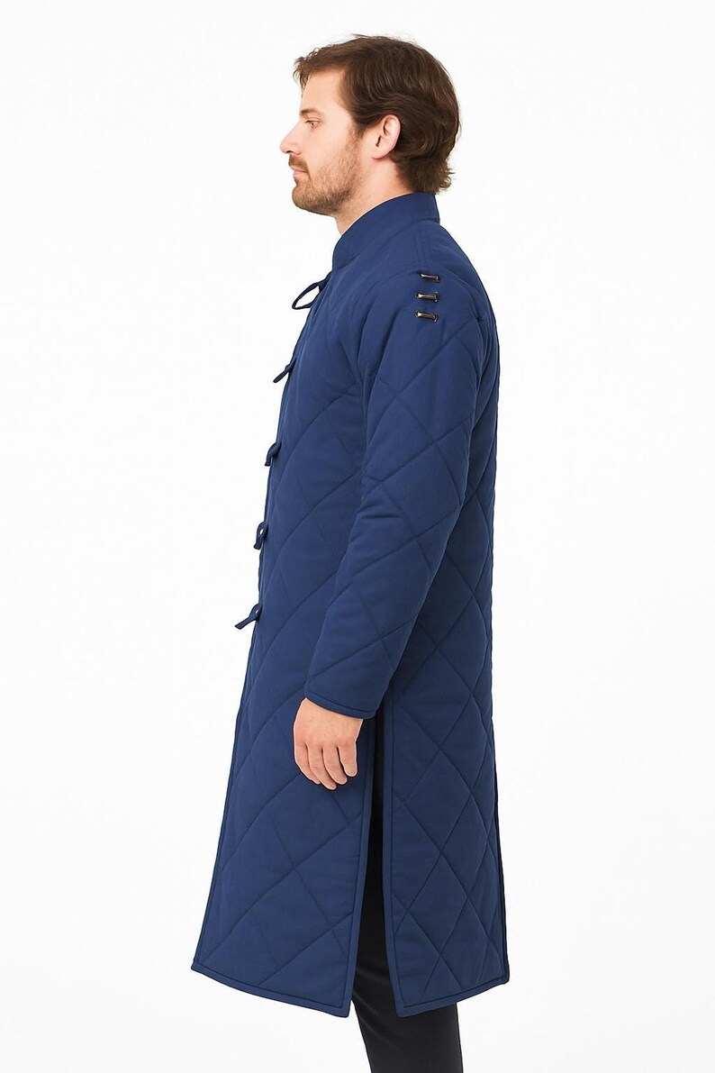 A male model wearing a long, blue quilted gambeson coat with a standing collar and front toggle closures, shown in profile against a plain white background.