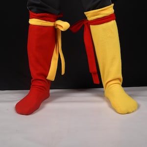 May include: A pair of colorful costume boots. One boot is red with a yellow ribbon tie, and the other is yellow with a red ribbon tie. The boots are knee-high and made of a soft material.