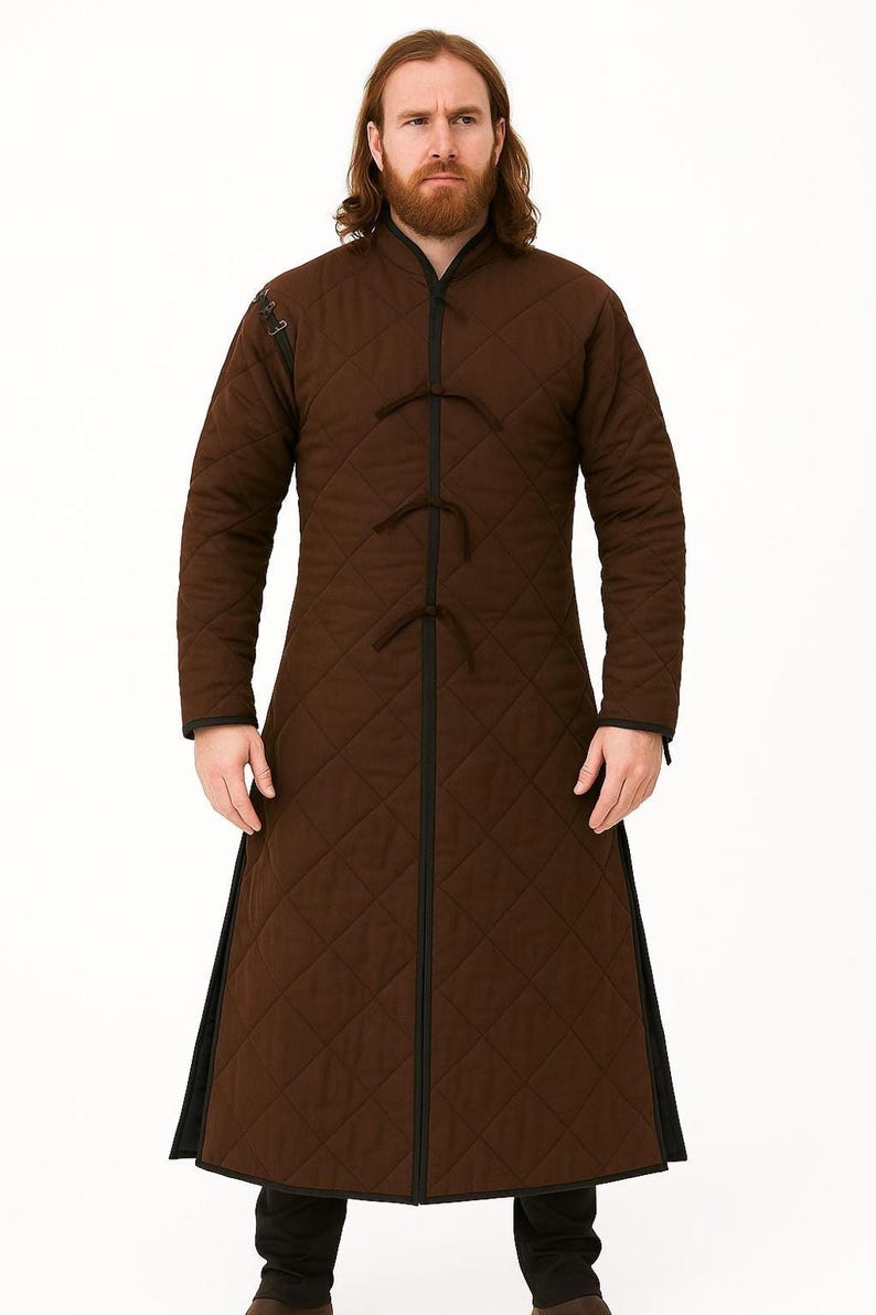 A man with a red beard wearing a long brown quilted gambeson coat with black trim and tie closures, standing against a white backdrop.