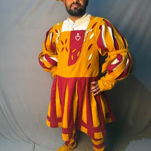 May include: A person wearing a Renaissance-style costume. The outfit features a yellow and red color scheme, with a hat, tunic, and leggings. The tunic has decorative cutouts and a heart-shaped emblem. The costume is made of a thick fabric.