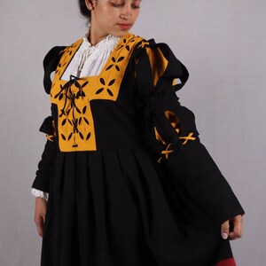 Pode incluir: Um vestido preto e amarelo em estilo medieval com mangas compridas. O vestido apresenta um decote quadrado com detalhes amarelos e uma camisa interior branca. As mangas t&ecirc;m la&ccedil;os decorativos e a saia tem acabamentos vermelhos e amarelos.