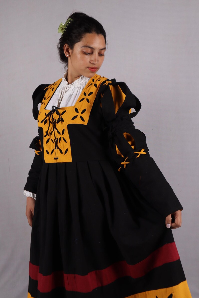 Pode incluir: Um vestido preto e amarelo em estilo medieval com mangas compridas. O vestido apresenta um decote quadrado com detalhes amarelos e uma camisa interior branca. As mangas t&ecirc;m la&ccedil;os decorativos e a saia tem acabamentos vermelhos e amarelos.