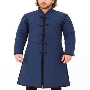 A cropped front view of a man wearing a blue quilted mid-length gambeson coat with black ties down the front.