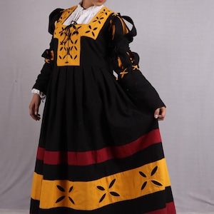 Pode incluir: Um vestido preto e amarelo em estilo renascentista com mangas compridas. O vestido apresenta um decote quadrado com uma camisa interior branca, detalhes amarelos e uma saia plissada com riscas vermelhas e amarelas. As mangas t&ecirc;m la&ccedil;os decorativos.