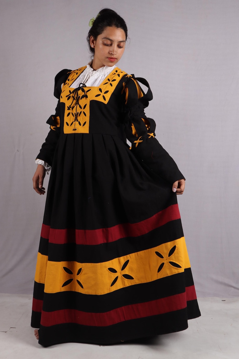 Pode incluir: Um vestido preto e amarelo em estilo renascentista com mangas compridas. O vestido apresenta um decote quadrado com uma camisa interior branca, detalhes amarelos e uma saia plissada com riscas vermelhas e amarelas. As mangas t&ecirc;m la&ccedil;os decorativos.
