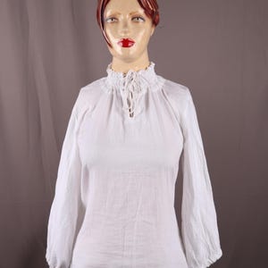 Pode incluir: Uma blusa branca de manga comprida, com gola alta com babados e decote em forma de fechadura. As mangas s&atilde;o franzidas nos punhos. O tecido parece ser linho leve ou algod&atilde;o. A blusa &eacute; exibida em um manequim.