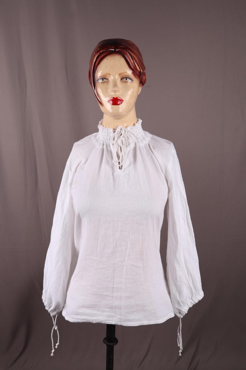 Pode incluir: Uma blusa branca de manga comprida, com gola alta com babados e decote em forma de fechadura. As mangas s&atilde;o franzidas nos punhos. O tecido parece ser linho leve ou algod&atilde;o. A blusa &eacute; exibida em um manequim.