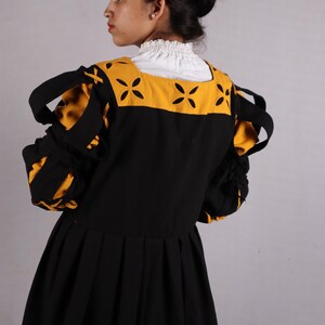 Pode incluir: Vestido preto com mangas amarelas e pretas. O vestido apresenta um decote quadrado com detalhes amarelos e uma gola branca com babados. As mangas t&ecirc;m detalhes listrados amarelos e pretos. O vestido tem uma saia plissada e uma faixa vermelha na bainha.