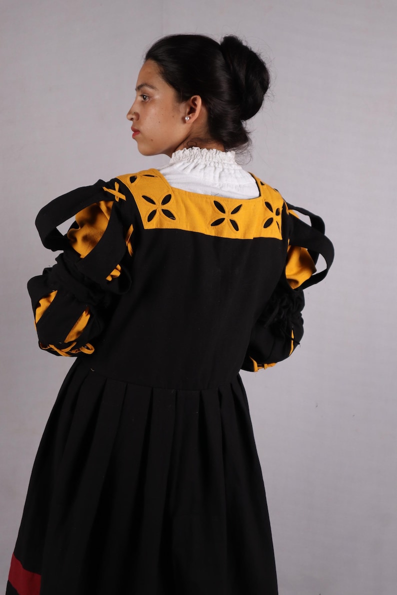 Pode incluir: Vestido preto com mangas amarelas e pretas. O vestido apresenta um decote quadrado com detalhes amarelos e uma gola branca com babados. As mangas t&ecirc;m detalhes listrados amarelos e pretos. O vestido tem uma saia plissada e uma faixa vermelha na bainha.