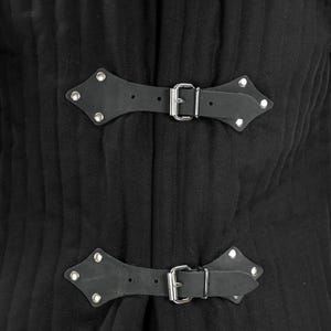 A close-up of two black leather strap closures with silver buckles and studs on a black quilted gambeson.