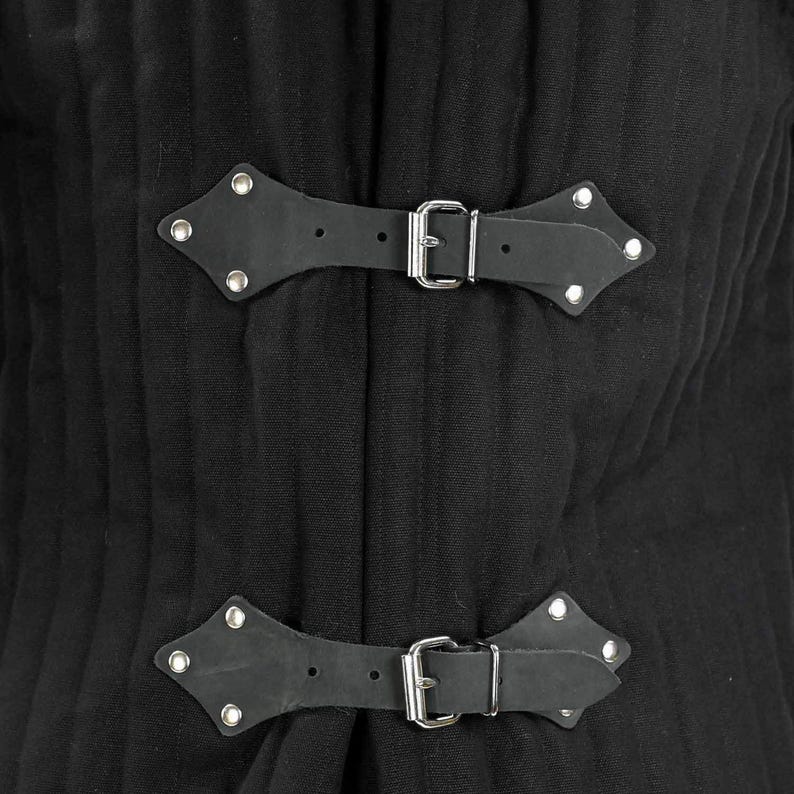 A close-up of two black leather strap closures with silver buckles and studs on a black quilted gambeson.